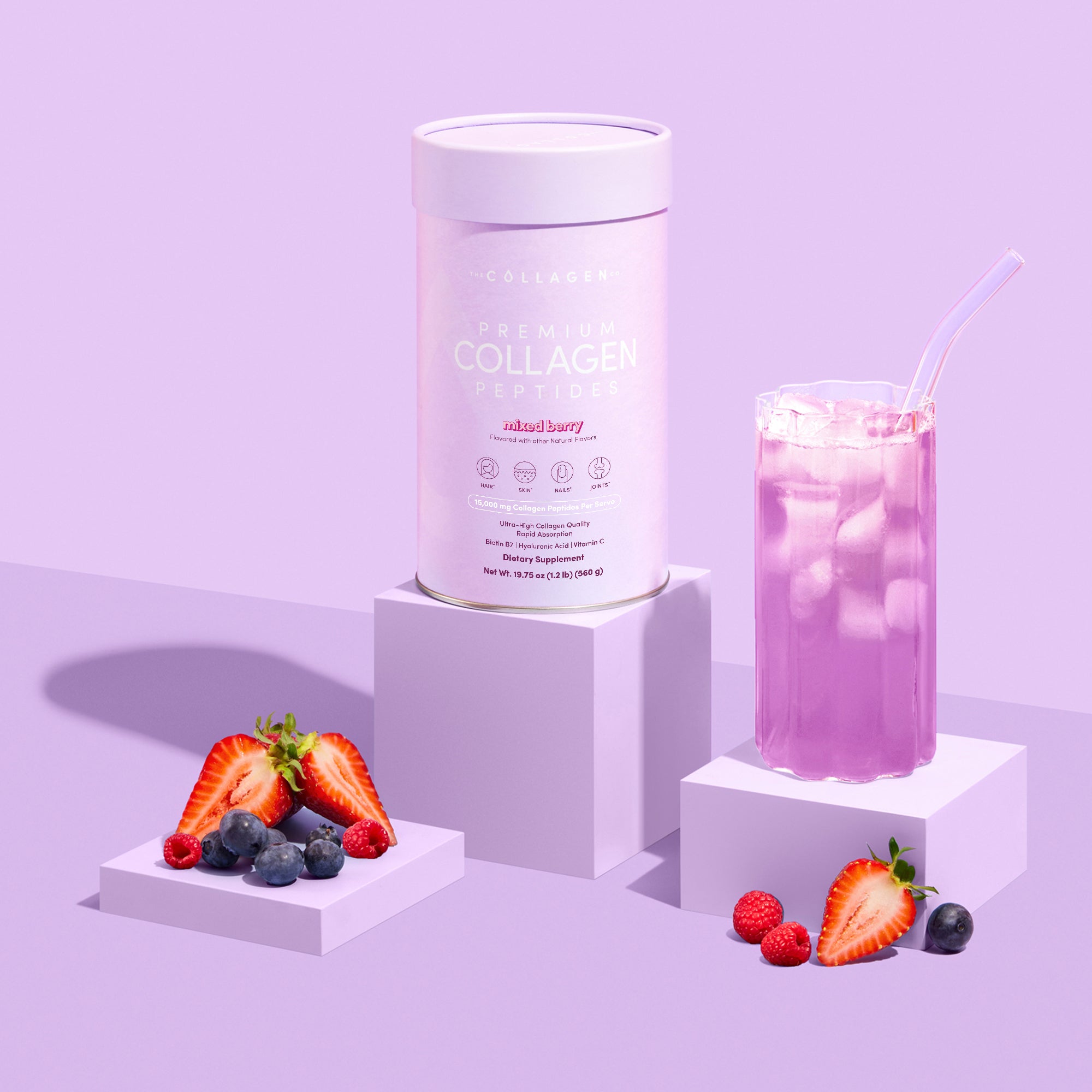 A tub of The Collagen Co. Mixed Berry Collagen Powder (560g) is placed between a glass of pink iced drink with a clear straw and a display of fresh berries, set on a pastel purple background.
