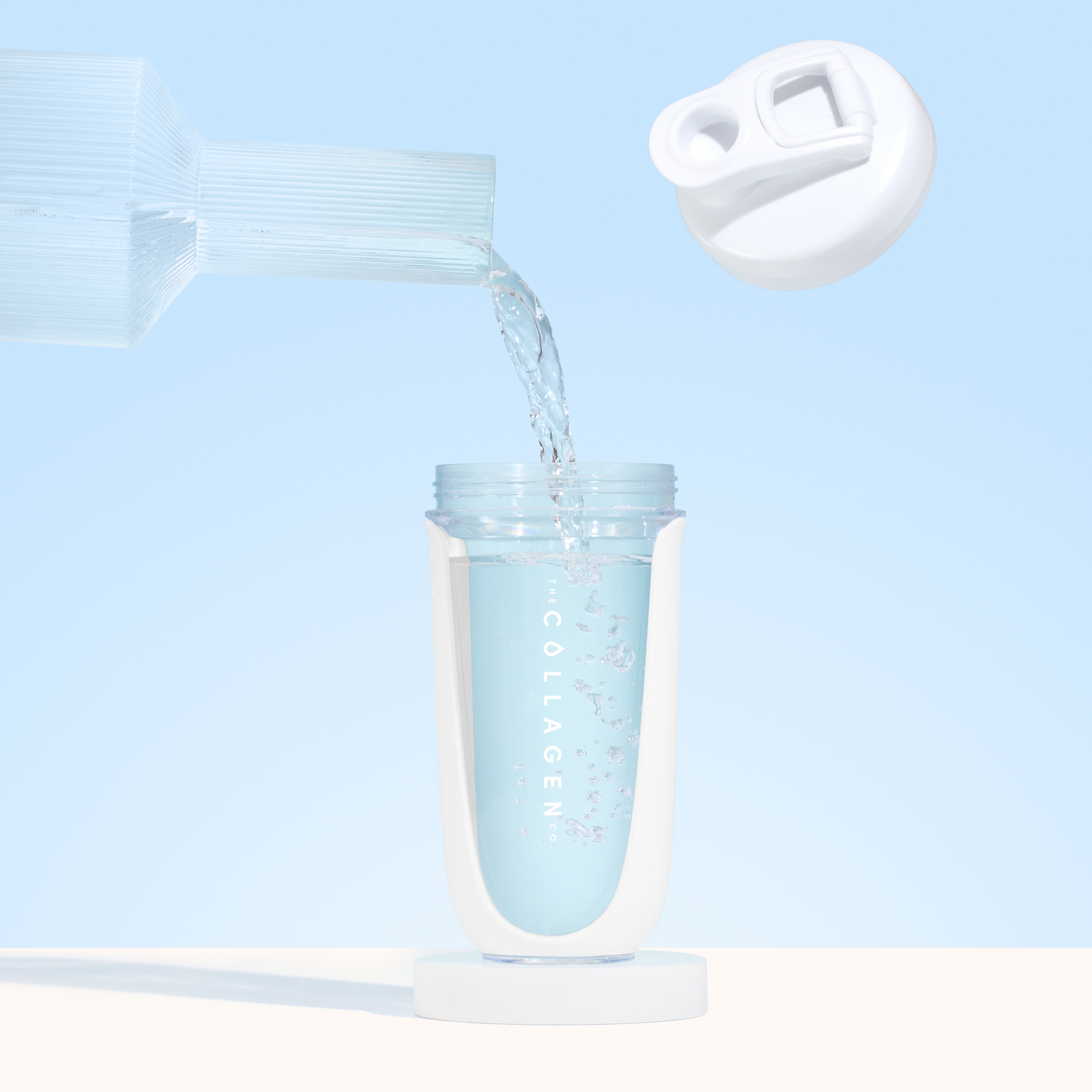 A close-up of water poured from a glass pitcher into The Collagen Co. White Collagen Protein Shaker - 600ml, labeled “Collagen,” with its BPA-free white lid open and floating above against a light blue background—a perfect fitness companion.