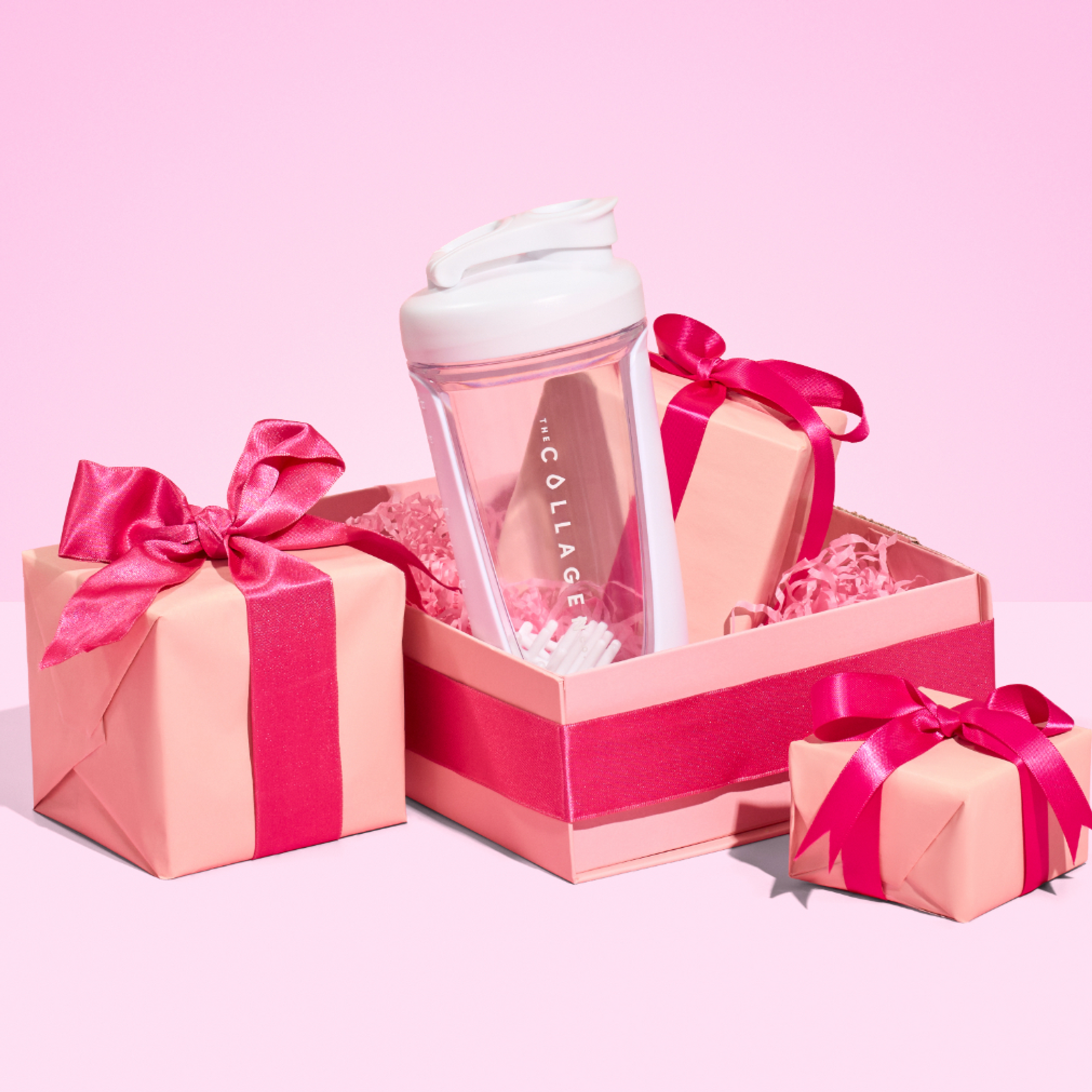 A vibrant pink gift box displays The Collagen Co. White Collagen Protein Shaker (600ml), with a pink accessory and two smaller ribbon-tied gift boxes on a vivid pink background—the perfect fitness companion for any occasion.