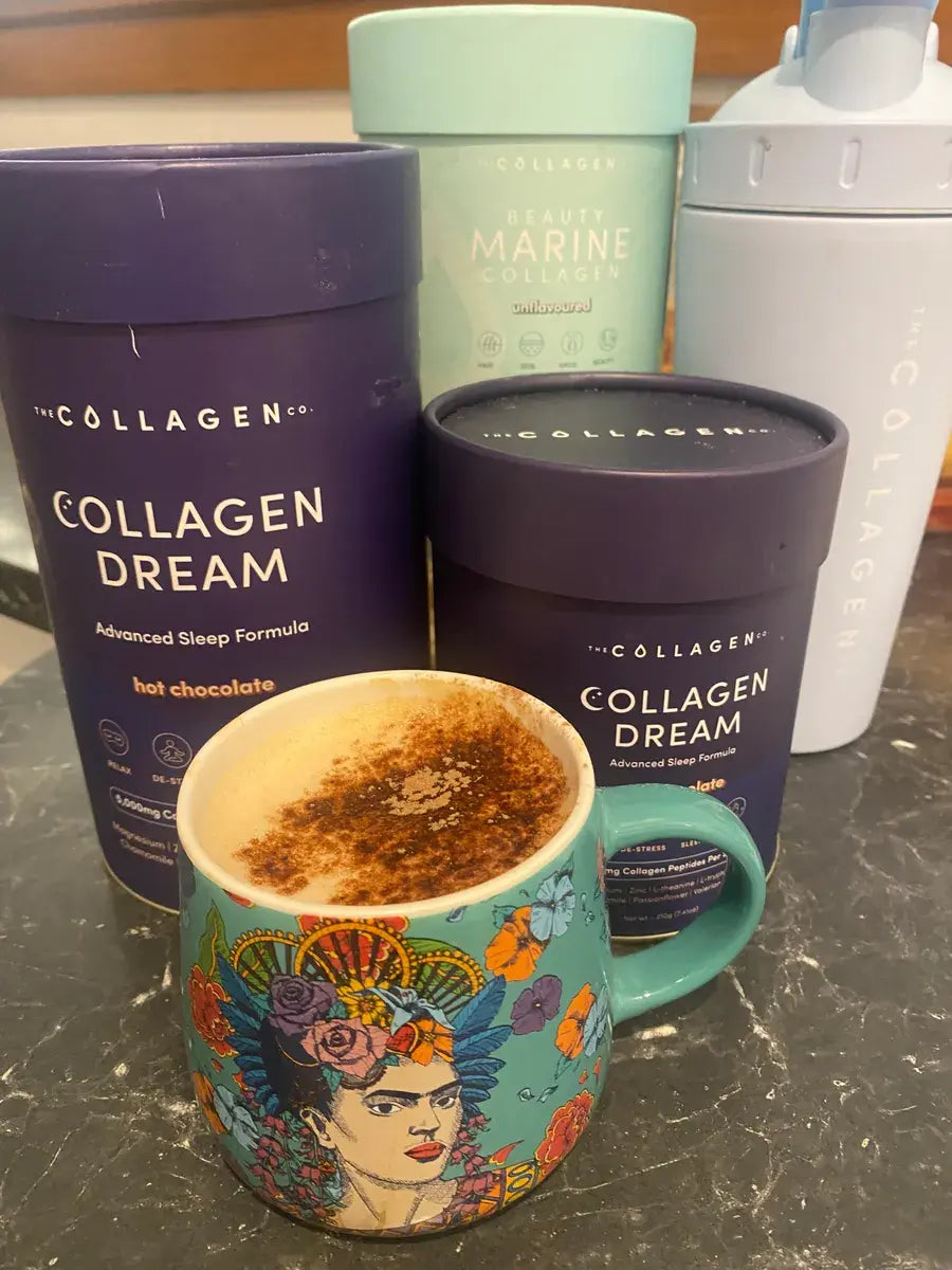 A colorful mug with a Frida Kahlo design sits on a dark countertop, filled with a frothy drink. Behind it are tall containers labeled Collagen Dream and Beauty Marine Collagen, plus a light blue shaker bottle.
