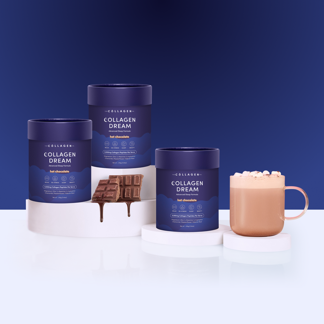 Three blue containers of the Custom Advanced Sleep Trio Bundle are displayed on white platforms, alongside a chocolate bar and a mug of hot chocolate with marshmallows—all set against a blue background, perfect for supporting skin health.