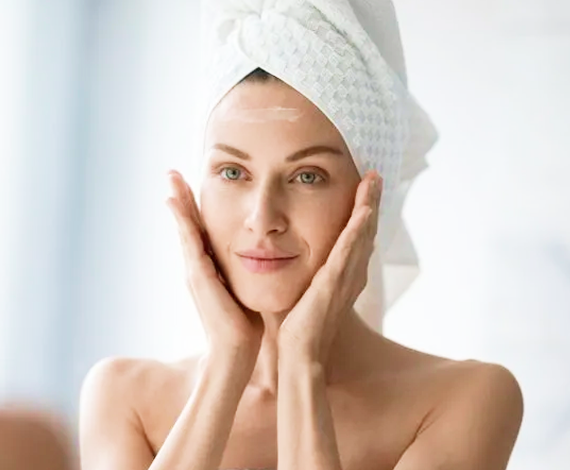 A woman with a towel wrapped around her head touches her face gently with both hands, smiling softly. She appears to be in a bathroom, possibly following a skincare routine.