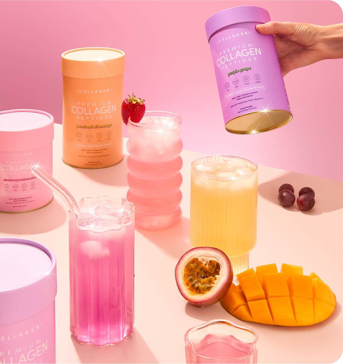 A colorful arrangement of collagen peptide containers, vibrant drinks, and fresh fruits including mango, passionfruit, grapes, and a strawberry on a glass, set on a pink background with a hand holding a lavender collagen container.