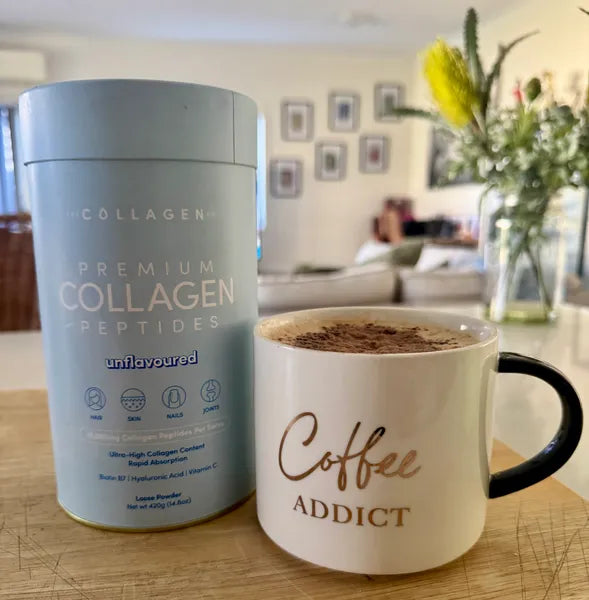 A container of premium collagen peptides sits next to a white mug labeled Coffee Addict, filled with coffee. The background shows a bright, modern living room with a vase of flowers and framed photos on the wall.
